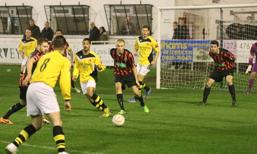Ein Sportplatz mit mehreren Männern, die Fußball spielen, ein Netz auf der rechten Seite, eine Tafel mit Text im Hintergrund und eine Gruppe von Menschen, die hinter der Tafel stehen und das Spiel beobachten, mit einer Wand und Fenstern im Hintergrund.