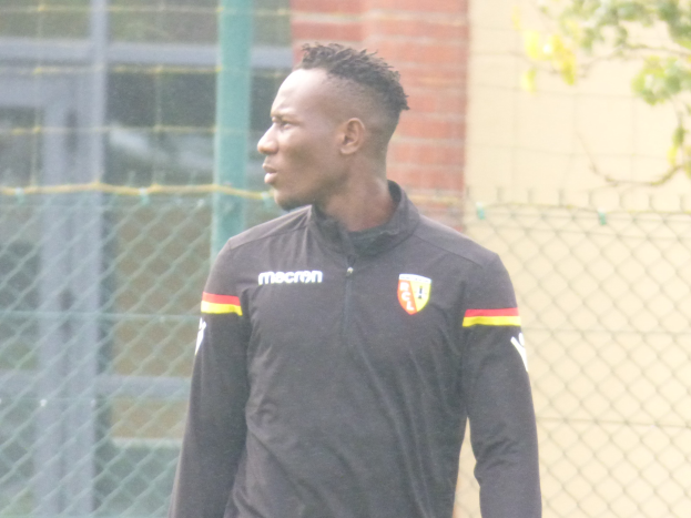 Ein Mann In Einer Schwarzen Jacke Steht Auf Einem Fussballfeld, Umgeben Von Einem Zaun Und Eines Baumes, Scheint Fokussiert Während Einer Trainingseinheit.