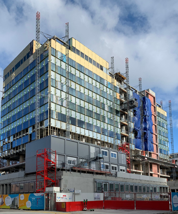 Großes Gebäude im Bau in einer Stadt mit Glasfenstern, Metallrahmen, Treppe, Zaun, Schild, Pfosten, Straßenlaterne, Bäumen und bewölktem Himmel.