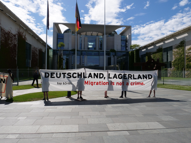 Eine Gruppe von Menschen mit einem Transparent, auf dem "Deutschland, Lagerland Migration ist kein Verbrechen" steht, vor einem Gebäude mit Fenstern, umgeben von Gras, einem Metallzaun, Pflanzen, Bäumen und Fahnen auf Stangen unter einem bewölkten Himmel.