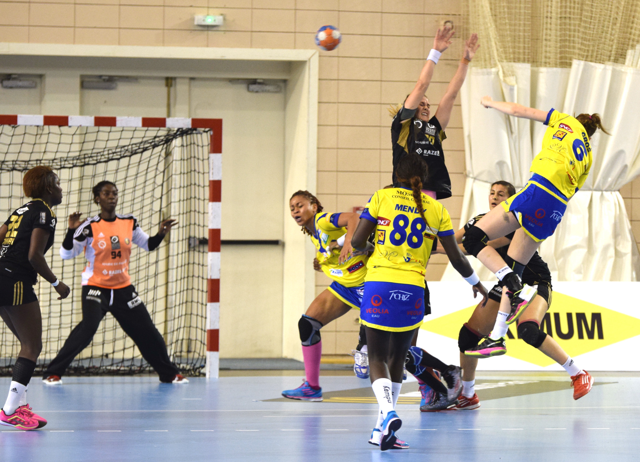 Eine Gruppe von Frauen, die Handball auf einem Feld spielen, mit einem Ball in der Luft, einem Torpfosten mit Netz, einer Fahne mit Text, einem Vorhang und einer Wand im Hintergrund.