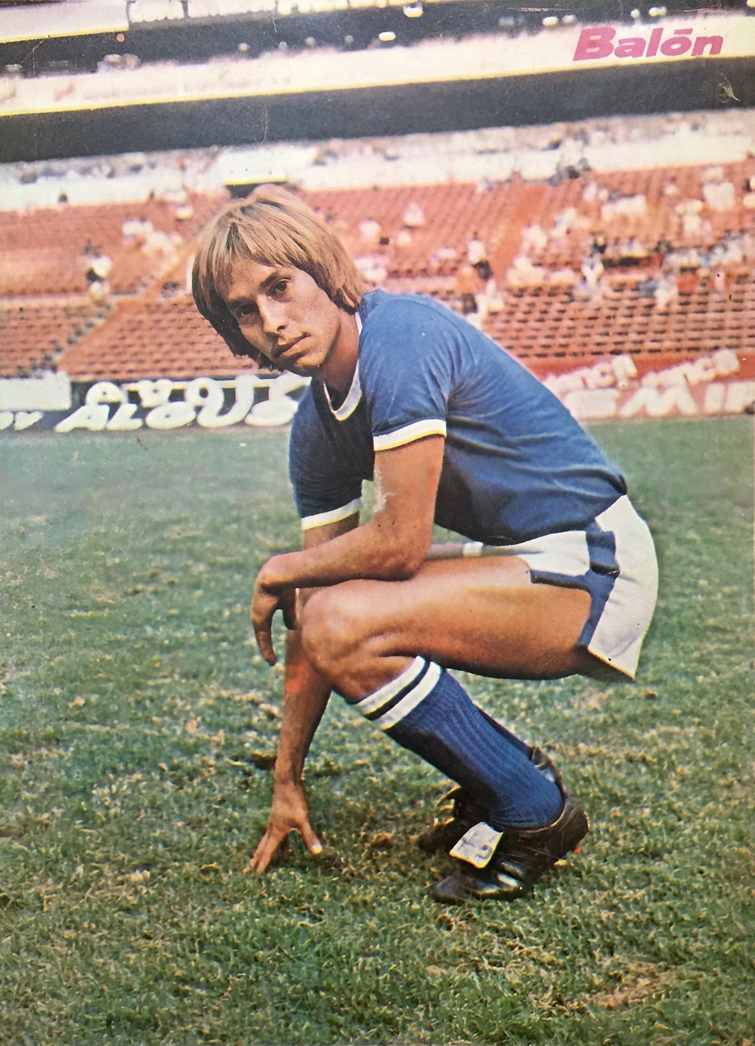 Ein Mann kniet auf einem Fußballfeld umgeben von Menschen auf Stühlen und Tafeln mit Text.