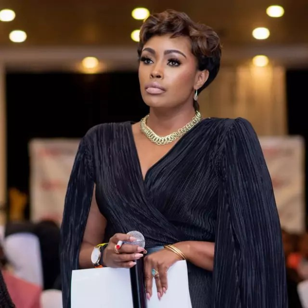 Eine Frau in einem schwarzen Kleid hält ein Mikrofon und ein Blatt Papier und spricht zu einem sitzenden Publikum unter Deckenbeleuchtung, während sie in eine Diskussion vertieft zu sein scheint.