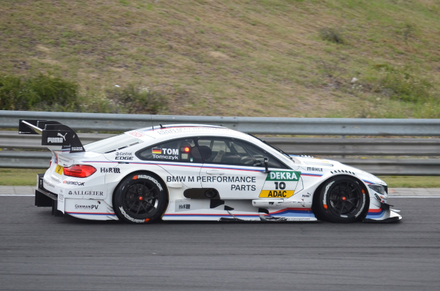 Ein BMW M4 DTM-Rennwagen auf einer Rennstrecke mit Text auf der Karosserie, umgeben von grünem Gras und einer Begrenzung im Hintergrund.