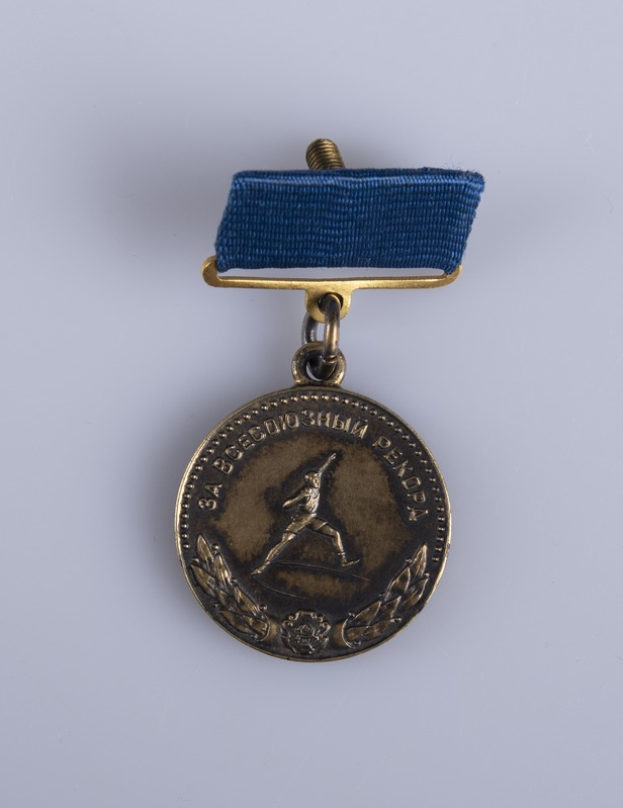 Eine Goldmedaille mit einer blauen Schleife, die eine laufende Person zeigt und likely mit Text beschriftet, der die Olympischen Spiele erwähnt.