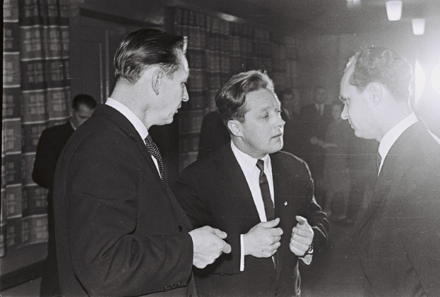 Drei Männer in Anzügen, die in einem schwach beleuchteten Raum mit Bücherregalen im Hintergrund diskutieren, in Schwarz-Weiß aufgenommen.