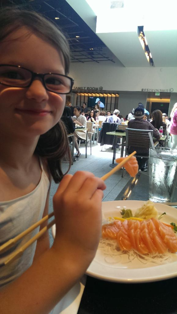 Ein Mädchen hält Stäbchen mit Essen vor einem Teller auf einem Restauranttisch, während andere Gäste an benachbarten Tischen im Hintergrund sitzen.
