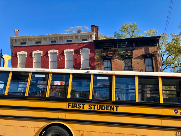 Gelber Schulbus mit der Aufschrift "First Student" vor einem roten Backsteingebäude mit einigen Menschen darin, Bäumen und einem klaren blauen Himmel im Hintergrund.