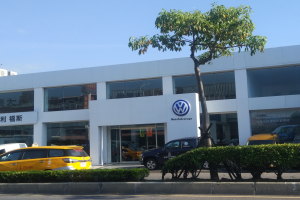 Ein VW-Autohaus mit parkenden Autos vorne, umgeben von Pflanzen und einem Baum, mit Glaswänden und Fenstern, unter einem sichtbaren Himmel.