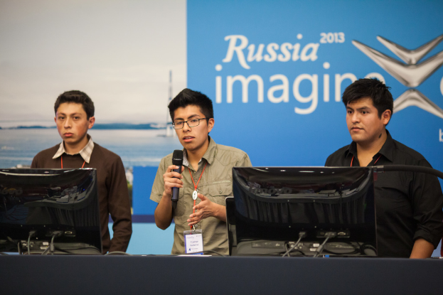 Drei Jungen stehen neben einem Schreibtisch mit zwei Computersystemen, einer spricht in ein Mikrofon, trägt ID-Karten mit roten Tags, vor einer Wand mit einer Werbetafel, die "Russland Imagine 2013" trägt.