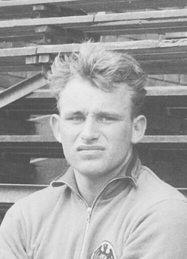 Schwarzes und weißees Foto eines Mannes in einer Jacke, der vor einem Haufen hölzerner Planken steht, der als legendärer deutscher Füßballer gilt.