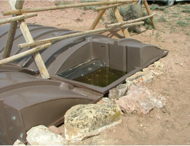 Kleiner Teich auf einem Schotterfeld, umgeben von Steinen und Holzstöcken, mit einem Fahrzeug und Pflanzen im Hintergrund und Text unten; der Teich sieht aus wie eine Klärgrube.