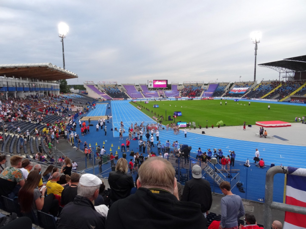 Ein großes Stadion voller Zuschauer bei einem Fußballspiel, mit Sitzplätzen und Stehplätzen, Geländern, einer Fahne, Flutlicht, einem Bildschirm, Bäumen und einem bewölkten Himmel.