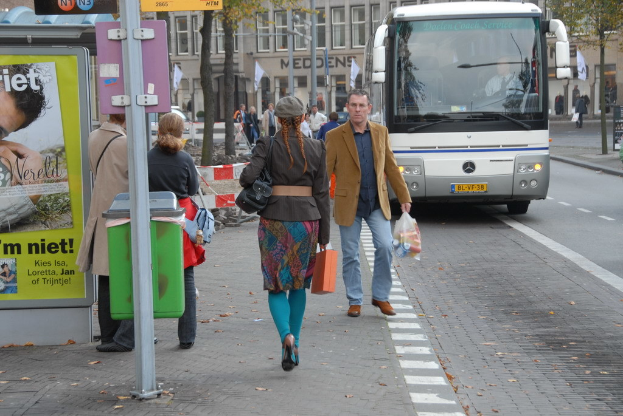 Eine Gruppe von Menschen, die auf einem Fußweg neben einem silbernen Bus laufen, mit einem braunen Gebäude mit Glasfenstern im Hintergrund und einer weißen und grünen Schildertafel auf der linken Seite.