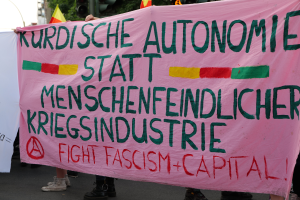 Eine Gruppe von Menschen steht auf einer Straße und hält ein pinkes Banner mit der Aufschrift "Kurdische Autonomie Statt Menschenfeindlicher Kriegsindustrie Fight Fascism Capital", mit Verkehrszeichen, Flaggen, Bäumen, Gebäuden und einem klaren blauen Himmel im Hintergrund.
