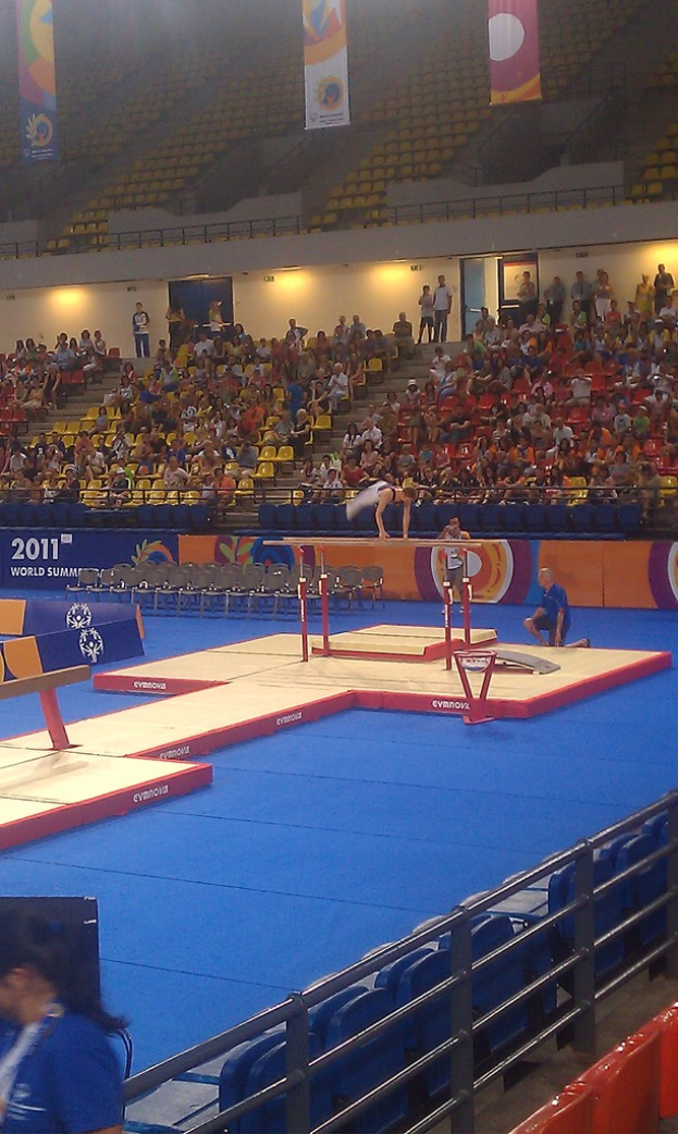 Eine Stadion-Szene mit Menschen auf Stühlen, andere im Hintergrund, Werbetafeln oben, Treppe in der Mitte und eine Person, die Gymnastik macht.