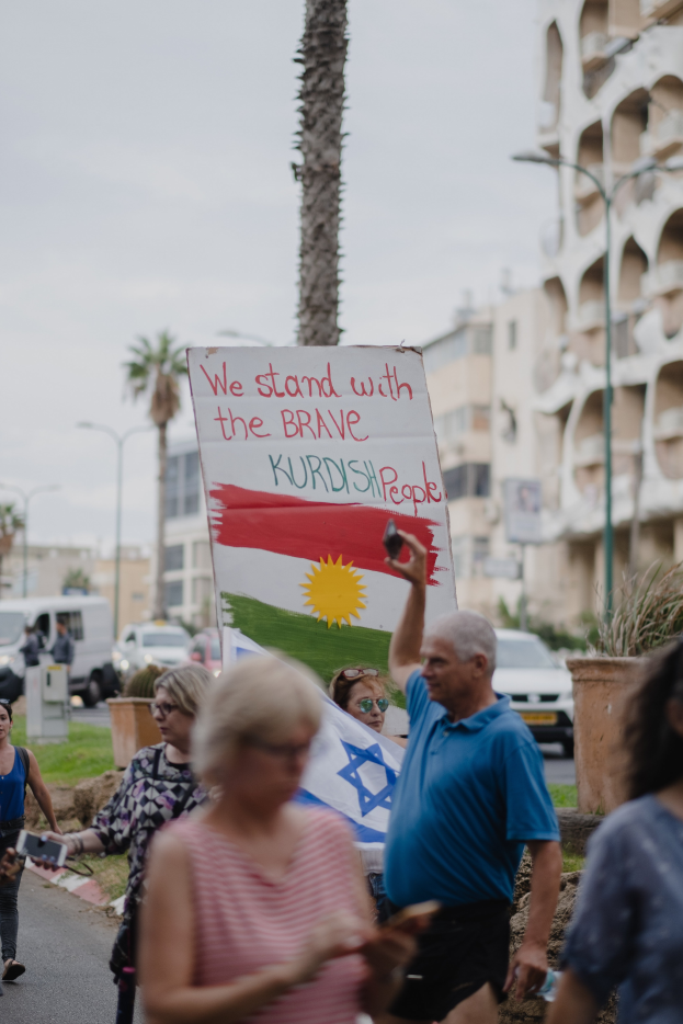 Eine Gruppe von Menschen marschiert nachts eine Straße entlang, hält ein Schild mit der Aufschrift "Wir stehen hinter den tapferen kurdischen Menschen", mit einem auf der Straße geparkten Van, Straßenlaternen, Bäumen, Gebäuden und einem klaren blauen Himmel im Hintergrund.
