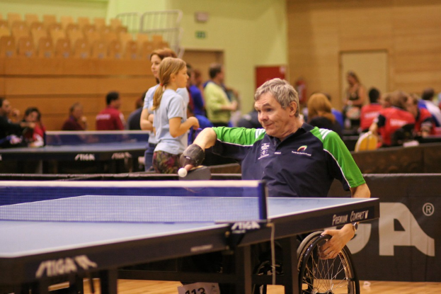 Ein Mann im Rollstuhl spielt Tischtennis, hält das Rad des Rollstuhls mit einer Hand und einen Schläger in der anderen, mit anderen Spielern und Tischen im Hintergrund.