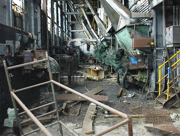 Ein altes, ungenutztes Fabrikinnere mit Maschinen, Schutt auf dem Boden, Fenstern und Gel├Ąndern auf beiden Seiten.