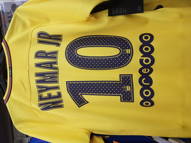Ein gelbes Fußballtrikot mit der Nummer 10, das Neymar Jr. von der Heimkit des PSG 2018-19 gehört, mit einem schwarzen Anhänger und Papieren und anderen Gegenständen im Hintergrund.