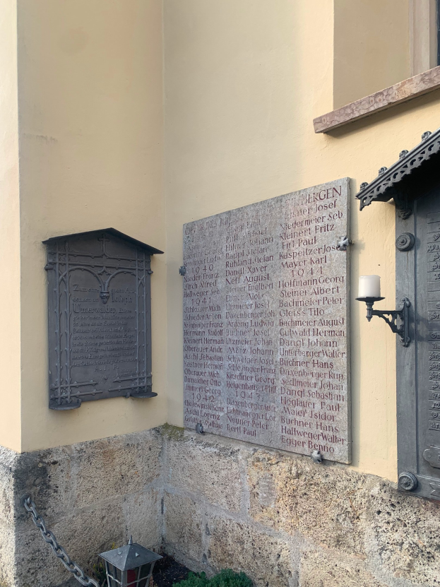 Ein Gebäude mit einer Holocaust-Gedenktafel, einem Fenster, einer Tür, einer Leuchte, einer Kette und umliegenden Pflanzen.