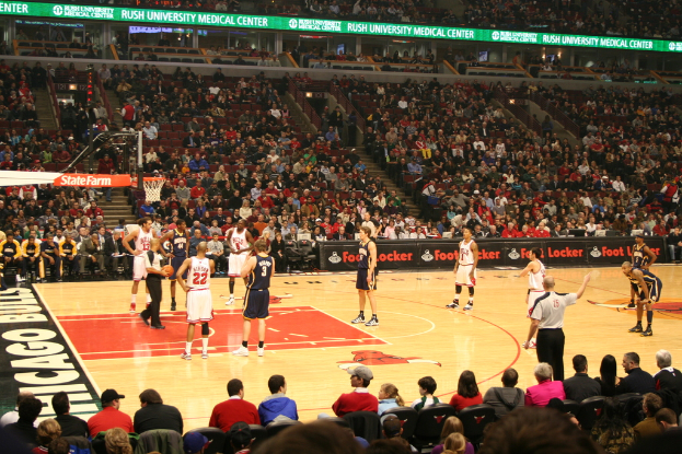 Eine Gruppe von Menschen in einer Arena, einige halten einen Basketball, andere schauen von den Rängen zu und Werbetafeln sind im Hintergrund sichtbar.