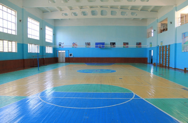 Große Turnhalle mit blau-weißem Boden, Basketballfeld, Netz, Stangen, einer Tür, Fenstern, Plakaten an den Wänden, einem Geländer und einem Dach.