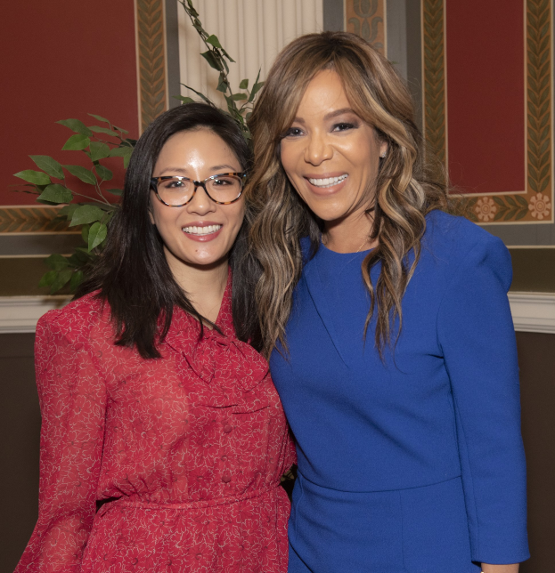 Zwei Frauen stehen nebeneinander und lächeln vor einer Wand; die Frau links trägt ein blaues Kleid, die Frau rechts trägt ein rotes Kleid und eine Brille, mit einer Pflanze und einem Vorhang im Hintergrund bei der 2019 Women in Film Pre-Oscar-Lunch-Veranstaltung.