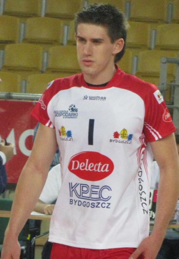 Ein Mann in rot-weißer Handballdressur steht auf einem Feld, umgeben von Zuschauern, mit einem Banner "Krasnoyarsk vs Bydgoszcz" im Hintergrund und entschlossener Miene.