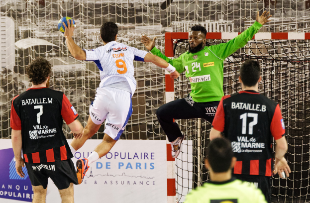 Eine Gruppe von Männern spielt Handball, einer springt und hält den Ball vor einem Torpfosten und einer Fahne mit der Aufschrift "Ligue 1 - Bordeaux vs Marseille".