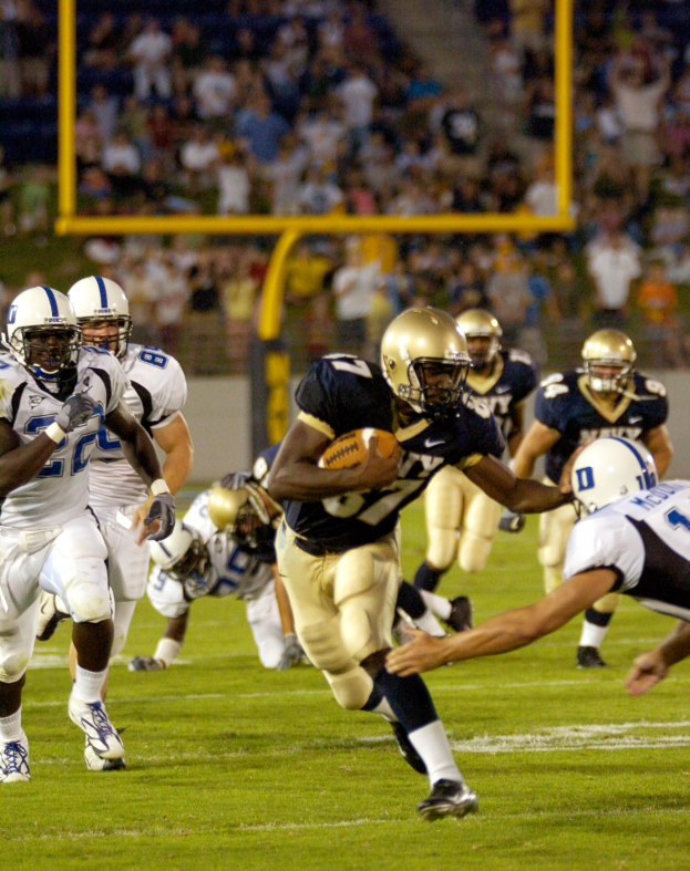 Ein Fußballspieler in einem Sportoutfit und Helm läuft mit dem Ball, während er von anderen Spielern umgeben ist, mit unscharfen Hintergrundelementen und Text am unteren Bildrand.