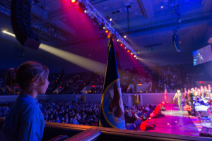 Ein junges Mädchen steht vor einer Menge auf einem Konzert, mit Musikern auf der Bühne, einer Flagge im Hintergrund und Lichtern und einem Bildschirm an der Decke, mit einem Geländer am unteren Bildrand.