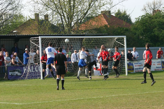 Spieler spielen Fussball auf einem Feld mit einem Tor, während Zuschauer dahinter stehen, mit Bäumen und Häusern im Hintergrund.