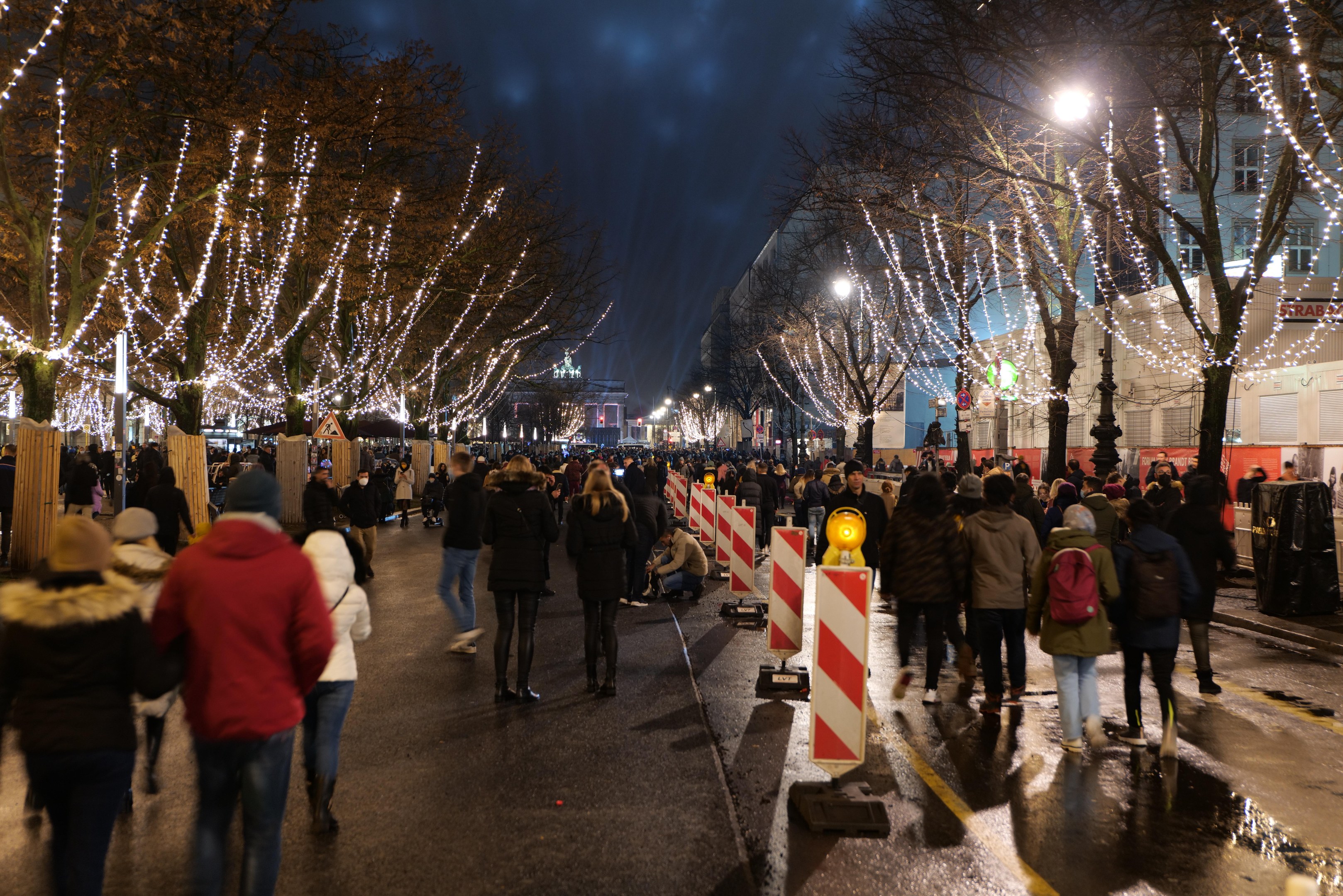 Eine Menschenmenge, die nachts eine Straße entlanggeht, beleuchtet von Weihnachtslichtern an Bäumen und Gebäuden unter einem bewölkten Himmel.