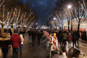 Eine Menschenmenge, die nachts eine Straße entlanggeht, beleuchtet von Weihnachtslichtern an Bäumen und Gebäuden unter einem bewölkten Himmel.