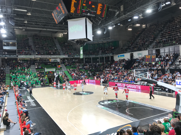 Ein Basketballspiel in einer großen Arena mit Zuschauern auf den Rängen, Anzeigetafeln mit "Asvel vs Galatasaray" und einer Decke mit Lichtern und einem Bildschirm.