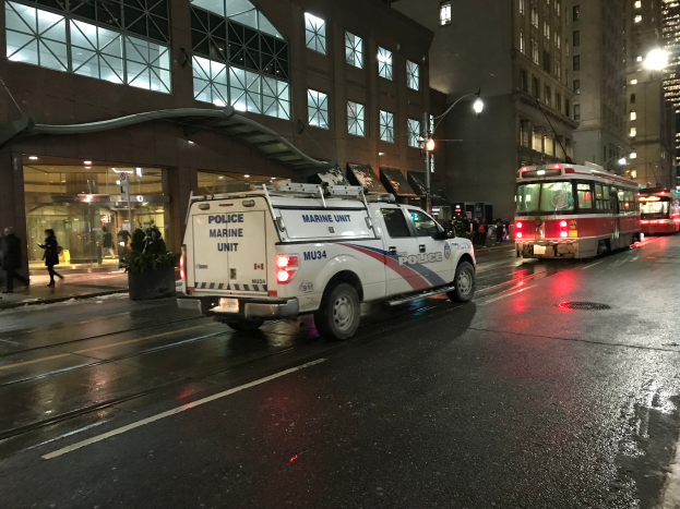 Ein Polizei-Lkw parkt nachts am Straßenrand einer Stadt, mit Fahrzeugen auf der Straße, Fußgängern auf dem Gehweg, Topfpflanzen, Straßenlaternen, Gebäuden mit Fenstern und einem bewölkten Himmel.