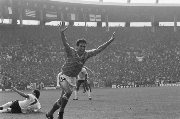 Ein Schwarz-Weiß-Foto eines Fußballspielers, der nach einem Tor jubelt, umgeben von Menschen auf dem Boden, mit Schildern, Fahnen und Pfählen im Hintergrund unter einem klaren Himmel.