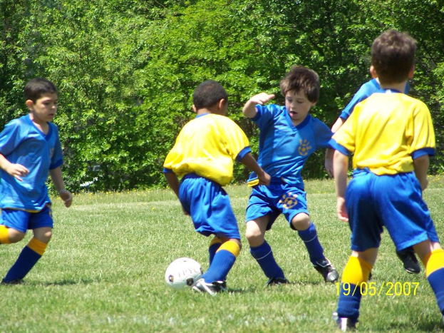 Kinder spielen Fußball auf einer grünen Wiese mit Bäumen im Hintergrund und einem Datum in der rechten unteren Ecke.