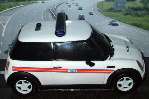 Ein weißer Polizeiwagen ist im Vordergrund geparkt, mit mehreren Autos, die sich auf der Straße dahinter bewegen, umgeben von Gras und Bäumen auf beiden Seiten, in einer ländlichen Umgebung.