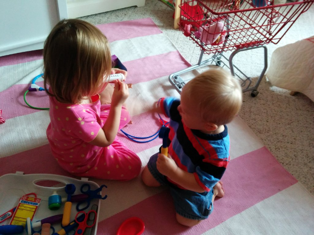 Kinder sitzen in der Mitte, ein Wagen rechts mit verschiedenen Gegenständen, ein weißes Objekt in der Nähe, ein Tablett unten links, eine Matte auf dem Boden mit einem Draht und einem roten Objekt darauf.