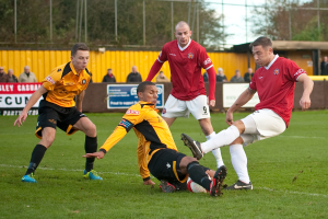 Spieler in blauen und roten Uniformen spielen ein Spiel auf einem grasbewachsenen Feld mit einem Ball, während Zuschauer außerhalb des Feldes jubeln, mit einem Baum und Himmel im Hintergrund.