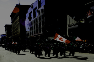 Eine Stadtansicht mit Gebäuden auf der rechten Seite, einer Straße unten, einer Gruppe von Menschen auf der Straße und einer weiteren Gruppe, die marschiert und Fahnen trägt, unter einem Himmel.