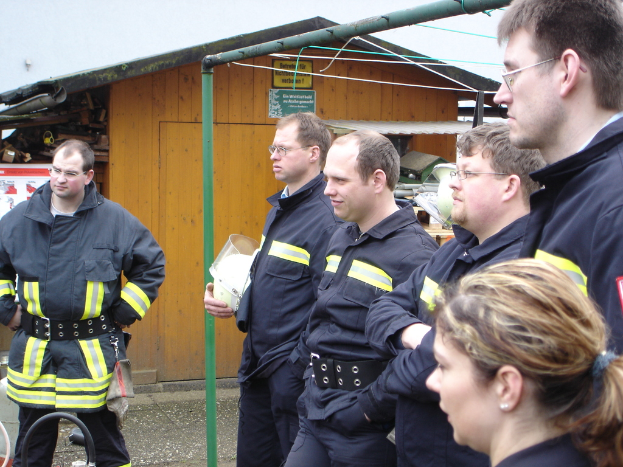 Eine Gruppe von Männern in blauen Feuerlöschuniformen, die nach links schauen, mit einem kleinen Holzhäuschen und Rohrrahmen im Hintergrund.