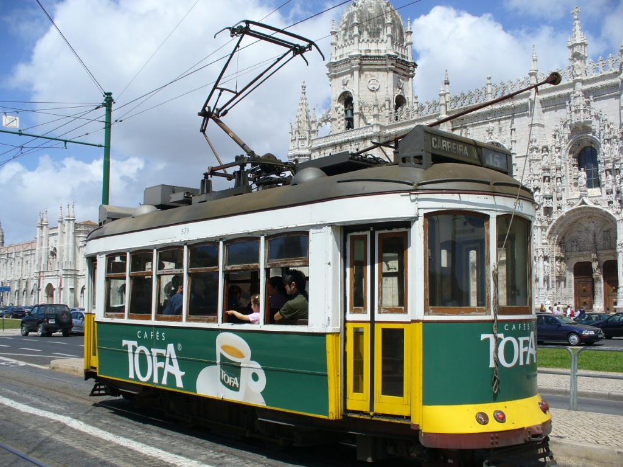 Eine Tram mit Menschen darin, Fahrzeuge auf der Straße, Gebäude, eine bewölkte Himmel, ein Strommast mit Drähten und ein Geländer in der Nähe.