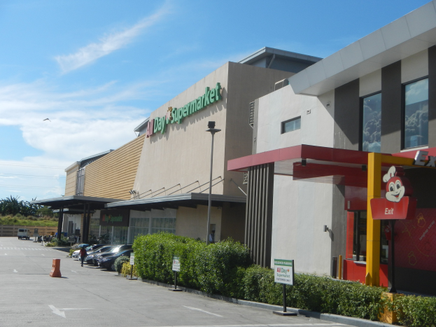 Außenansicht eines Supermarkts mit einem Parkplatz davor, umgeben von Gebäuden und Straßeninfrastruktur, mit Fahrzeugen, Absperrbaken und Grünflächen unter einem bewölkten Himmel.