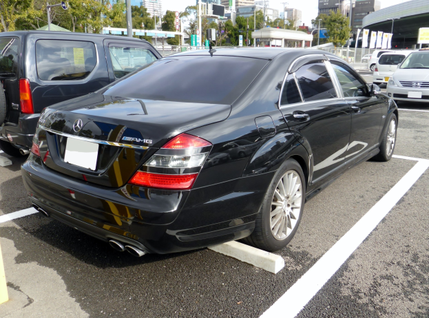 Schwarzer Mercedes S-Klasse in einer Parklöcke mit anderen Autos umgeben von Gebäuden, Bäumen, Masten, Werbetafeln und einem klaren blauen Himmel.