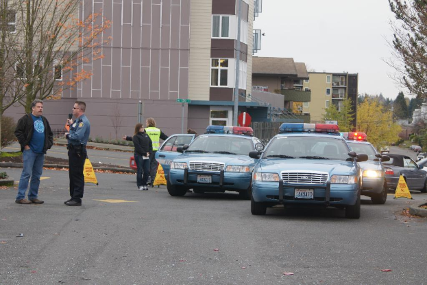 Autos auf einer Straße mit vier Personen in der Nähe, Gebäude mit Fenstern im Hintergrund, Bäume und ein Warndreieck.