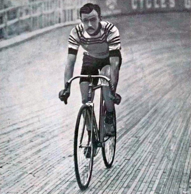Altes Schwarz-Weiß-Foto eines Mannes, der auf einer Rennbahn Fahrrad fährt, mit einem Zaun und einer Tafel im Hintergrund.