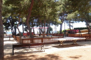 Eine Parkszene mit einem zentralen Tischtennis-Tisch, umgeben von Bänken, einem Zaun, Schaukeln, Pfosten, Pflanzen und Bäumen, mit einem Wasserbecken und einem klaren blauen Himmel im Hintergrund.